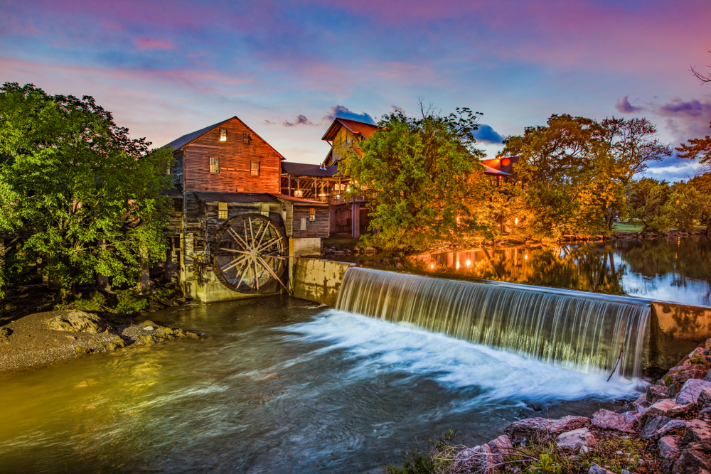 The old grist mill in Pigeon Forge, one of the best small towns in Tennessee. There is a large man made waterfall and trees and rocks surrounding the river. The sun is setting and the sky is purple and blue. 