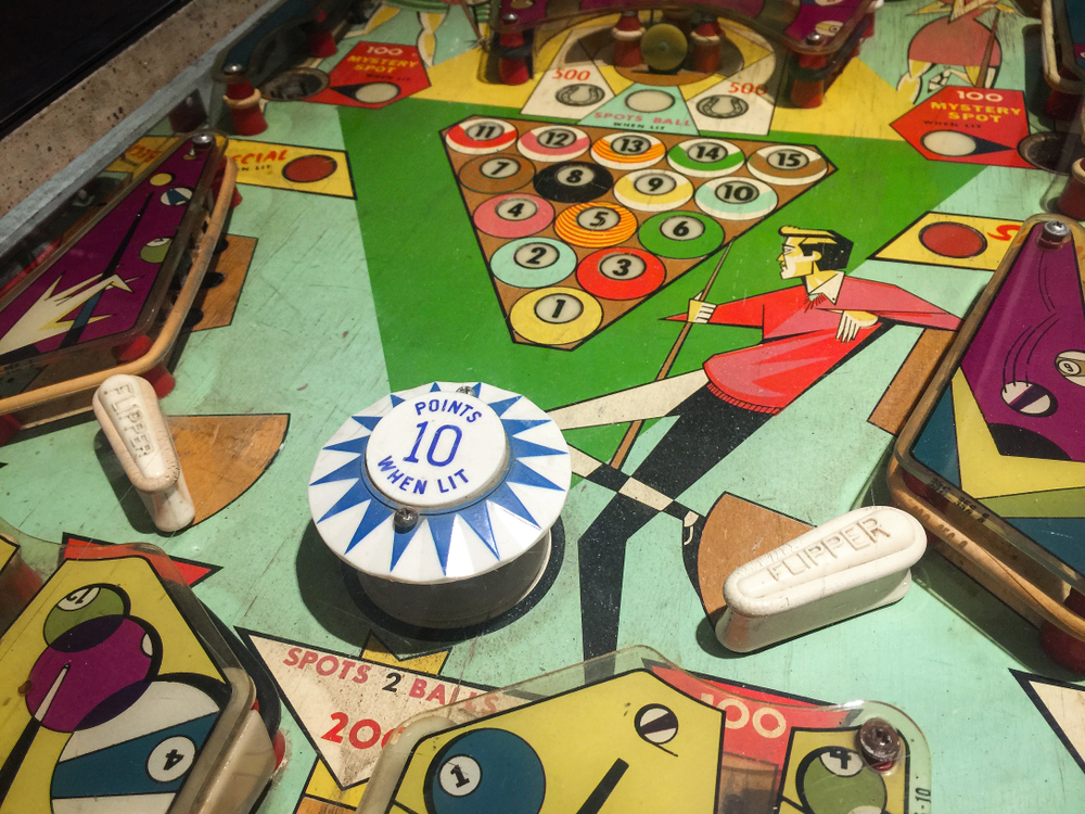 A close up image of an antique pinball machine. It has decorations that are themed around pool. There is a guy holding a pool stick, pool balls, and more. 