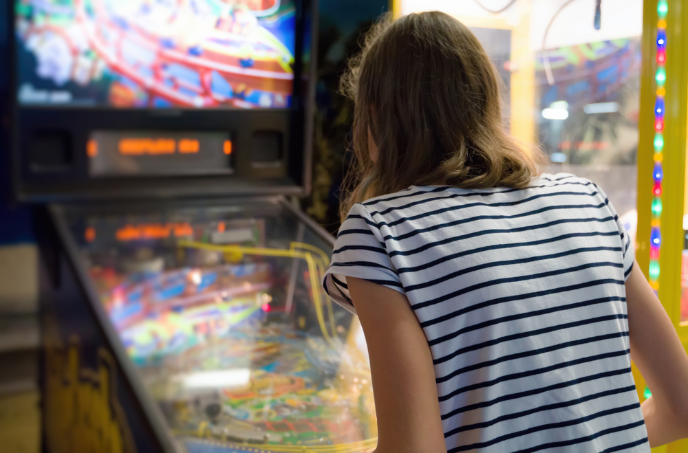 A person with shoulder length hair wearing a striped shirt. They are bent over and playing a pinball machine.