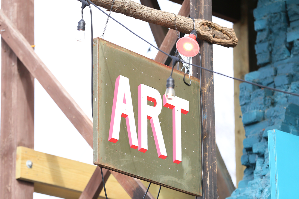 A sign on a wooden pole that reads 'ART' in block letters that are white, red, and pink. The sign is an army green color. You can see a string of lights near it and a brick building behind it.