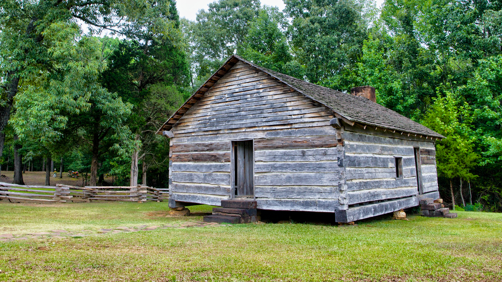 An old wooden cabin in a grassy area. Behind the old cabin is a line of trees with green leaves and an old wooden fence that is made of stacked logs. 