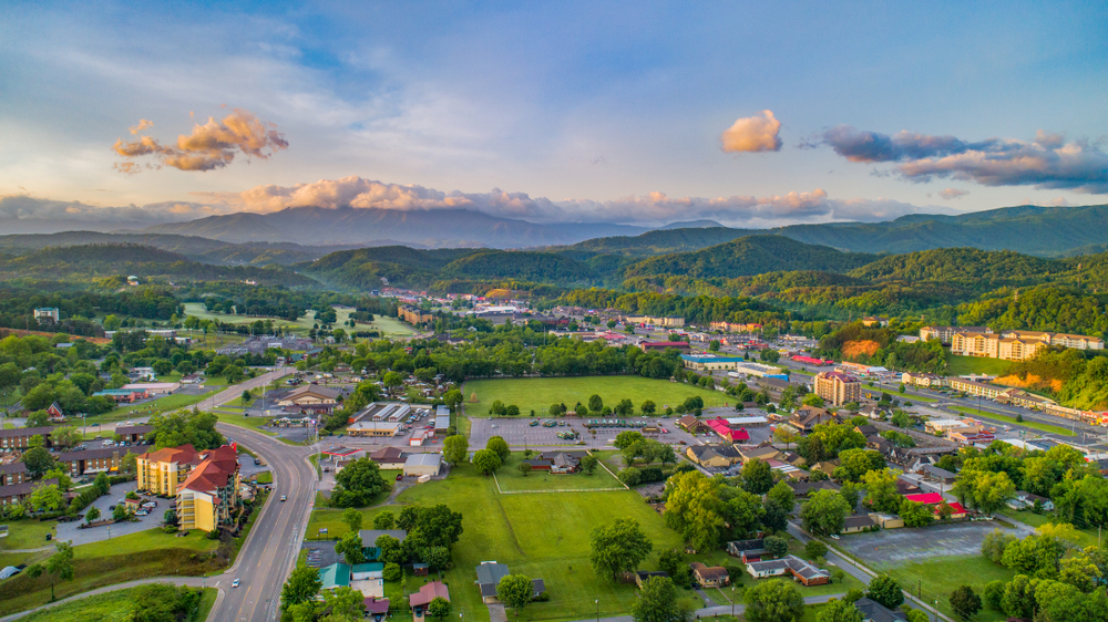 An aerial view of one of the small towns in Tennessee. You can see mountains in the distance and smaller buildings in the valley. There are large green areas and lots of green trees. 