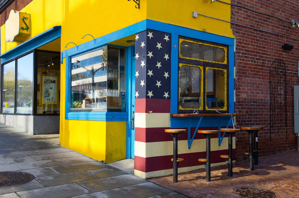 A building with the front painted bright yellow with blue trim. On the side there is an American flag painted on the building, the rest is brick. There is a counter at a window you can sit at and there are stools. 