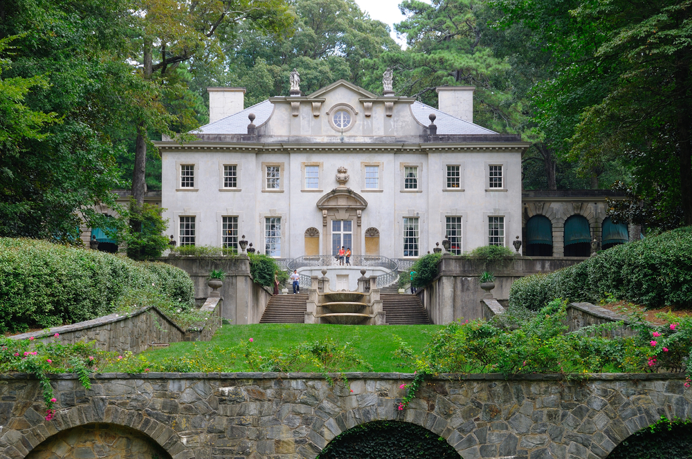 A Greek Revival mansion in Georgia that is a popular spot for filming. It is a cream color with lots of square windows and a stretch of green grass surrounded by shrubs in front of it. Behind it is dense trees.