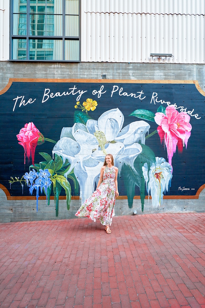 A woman in a floral maxi sundress standing in front of a black and floral mural in Georgia. The mural is on the side of an old industrial building. 