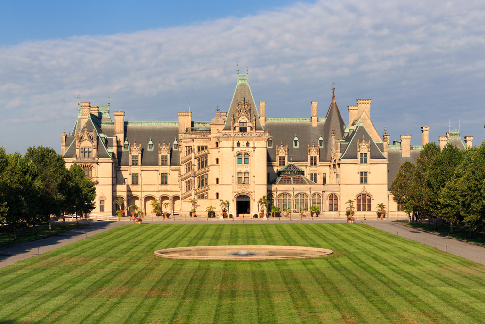 The front exterior of the Biltmore Estate. It is a large French-Gothic style house with tan walls, a grey roof, and blue green trim. In front of it is a large grassy area with a fountain in the middle. Around the grassy area and in front of the house is a paved walkway.