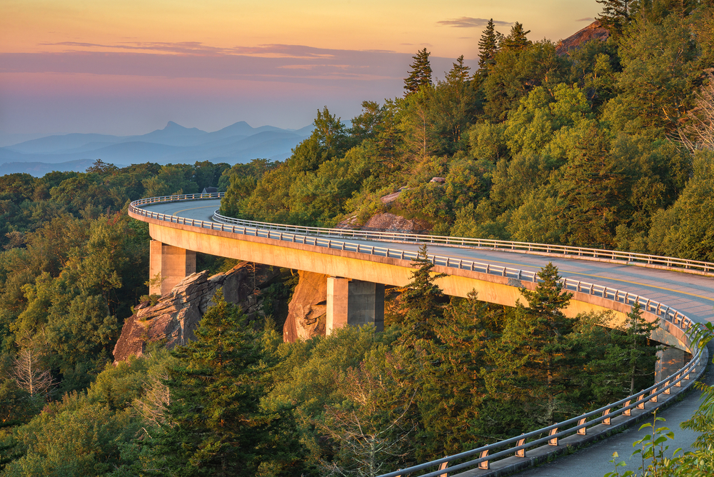 The Linn Cove Viaduct area of the Blue Ridge Parkway. It is a winding road that is lifted off of the rocky cliffside from supports underneath. The cliff next to it is covered in tall trees with green leaves. In the distance you can see other mountains silhouetted.