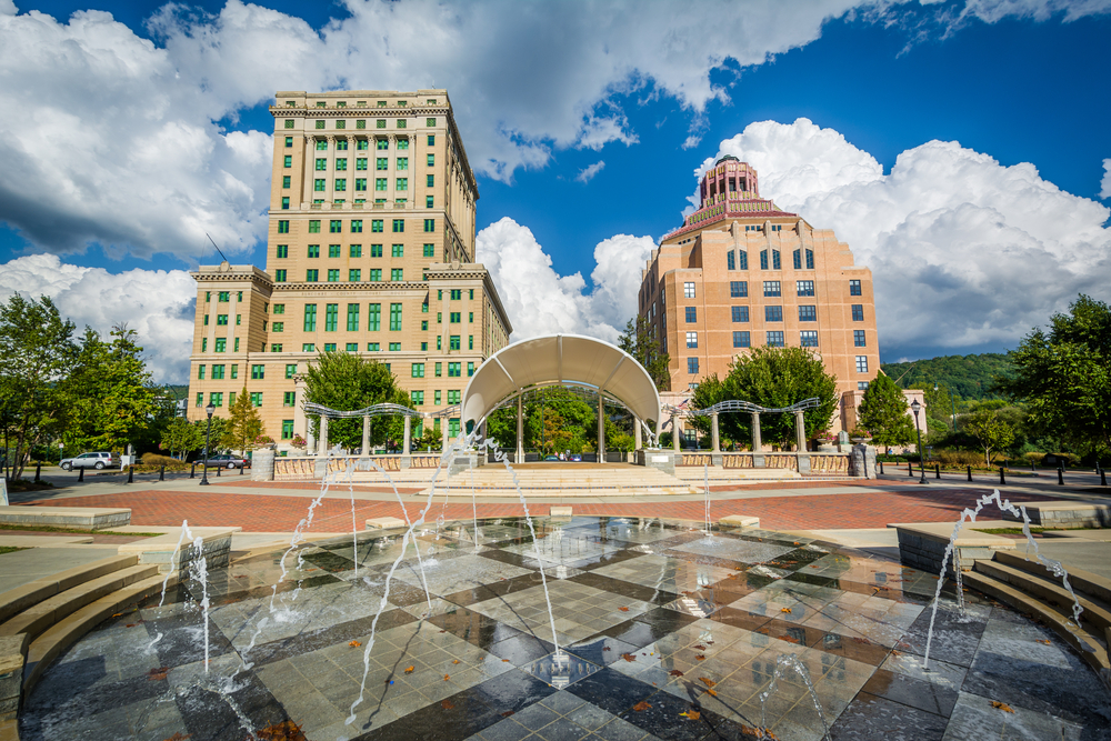 A courtyard in front of two Art Deco style buildings. In the courtyard there is a water feature with marble tiles and water shooting out. There is also a large brick paved area and what looks like a covered stage. 