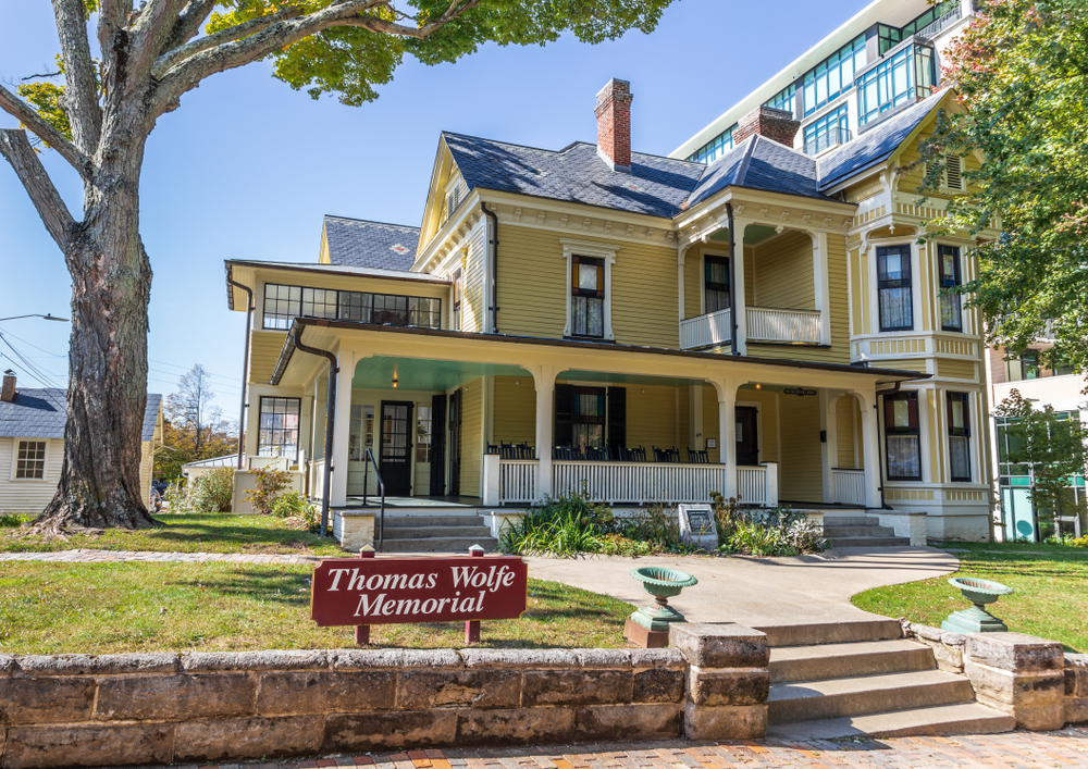 The exterior of a yellow Victorian house. It has a large front porch with rocking chairs on it, windows with black trim, and a small walkway leading to the front steps. There is a small lawn and a red sign that says 'Thomas Wolfe Memorial'. 