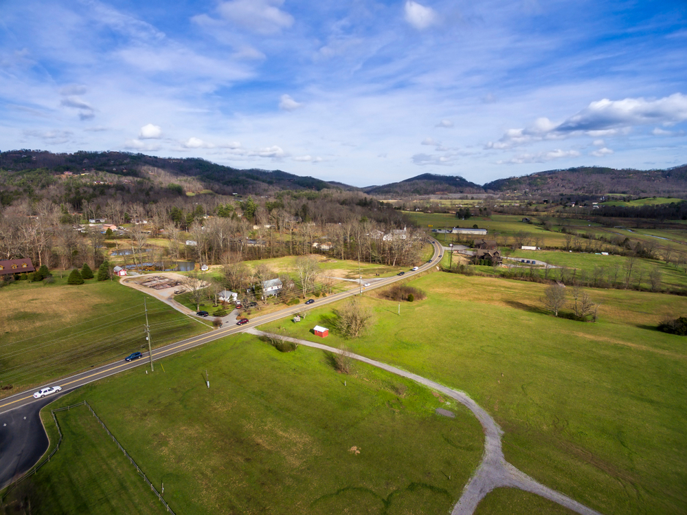 An aerial view of the small town of Townsend, one of the cutest small towns in Tennessee. It has a lot of green fields, smaller buildings, and you can see cars on the road. The trees don't have any leaves and you can see mountains in the distance. 