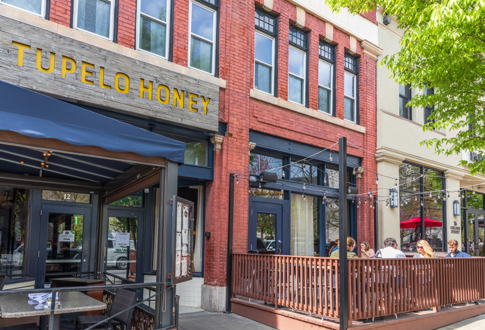 The exterior of a brick building in a row of buildings. Above it is a sign that says "Tupelo Honey' in yellow writing on a wooden background. There is a black tent with seating under it. There is also a patio with string lights over it where you can see people sitting and eating. 