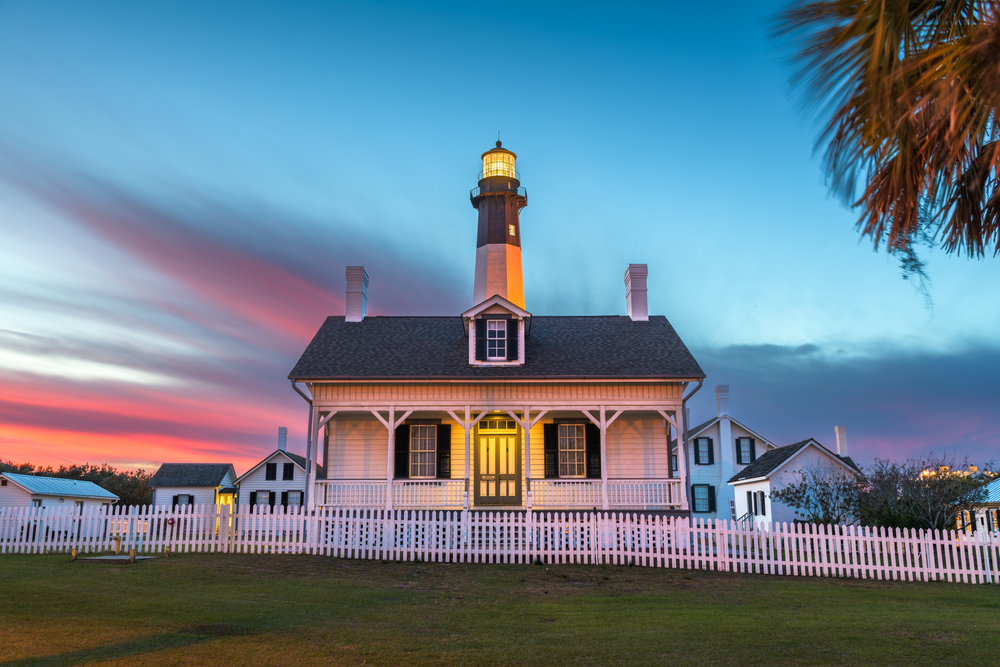 The lighthouse on Tybee Island. It is a small white house with black shutters and a front porch. Behind it is the striped black and white light tower. It is sunset and the sky is pink, blue, purple, and orange.