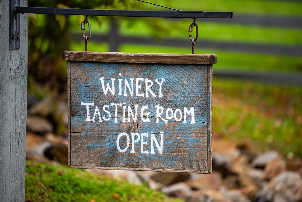 A sign hanging from a wooden post that say 'Winery Tasting Room Open'. It is a wood sign that has distressed blue painted on it. The words are white.