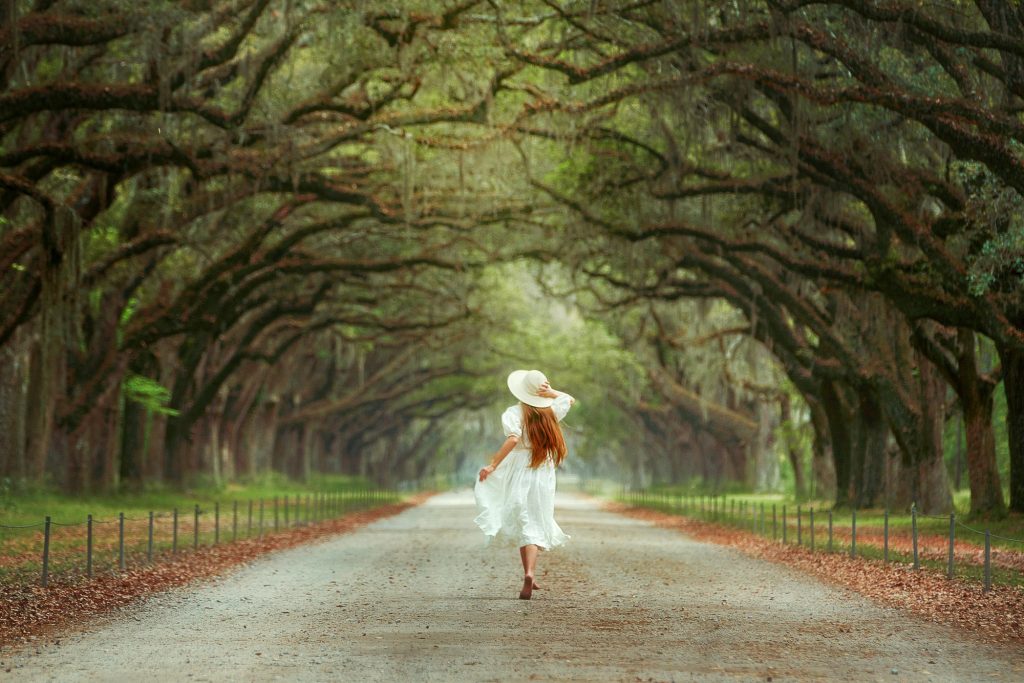 A woman in a white dress and sun hat with long hair running barefoot down a dirt road. On either side of the road there are live oak trees that create a tunnel. The trees are covered in Spanish moss.