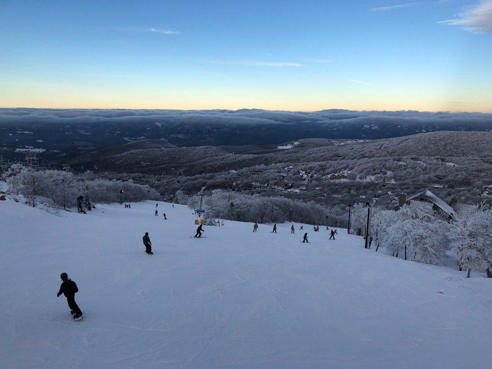 A picture snowboarders going down the side of a snowy bank at Beech Mountain Ski Resort with rolling hills and mountains in the distance.