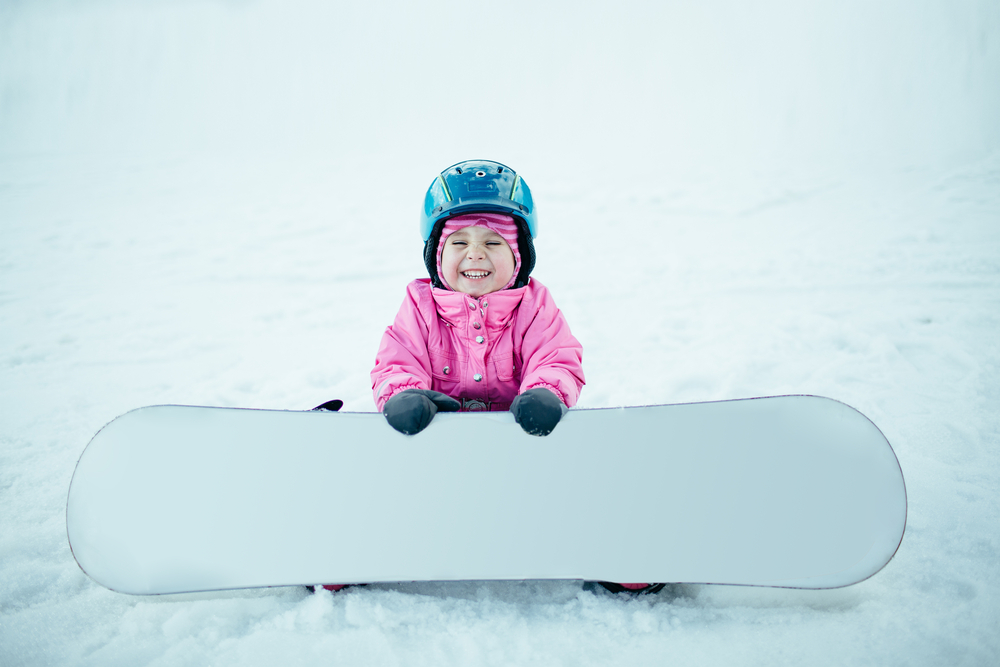 A photo of a child smiling who is wearing a blue helmet, sitting on snow, and holding a white snowboard.