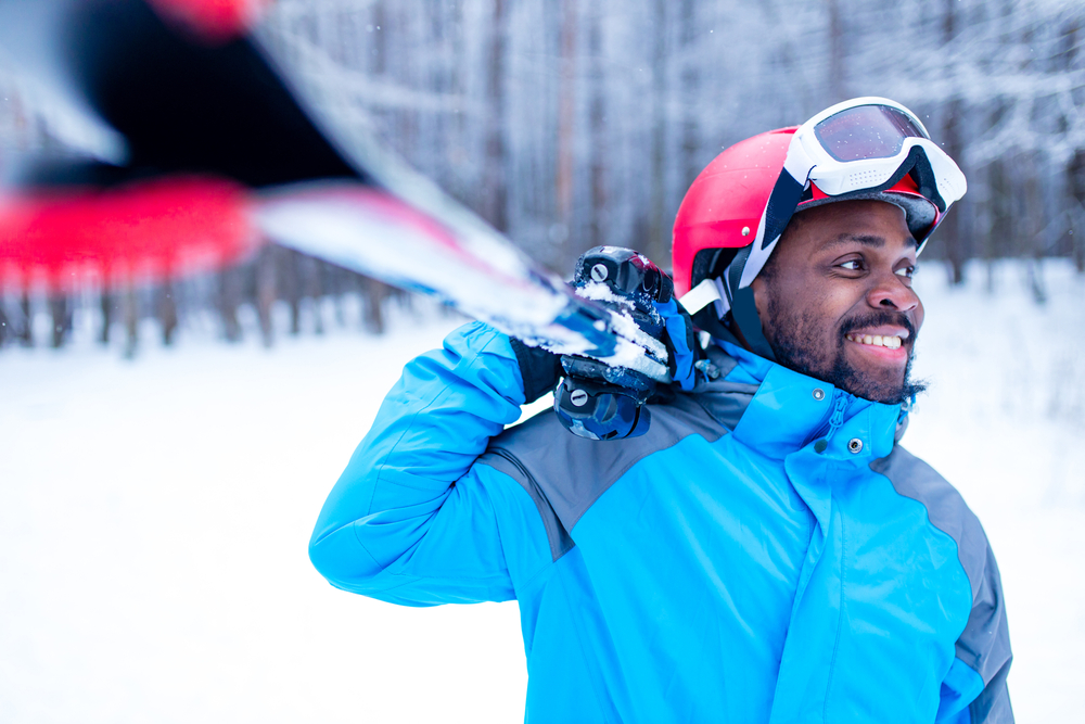 A photo of a man smiling in a blue coat holding a pair of skis on a snowy day.