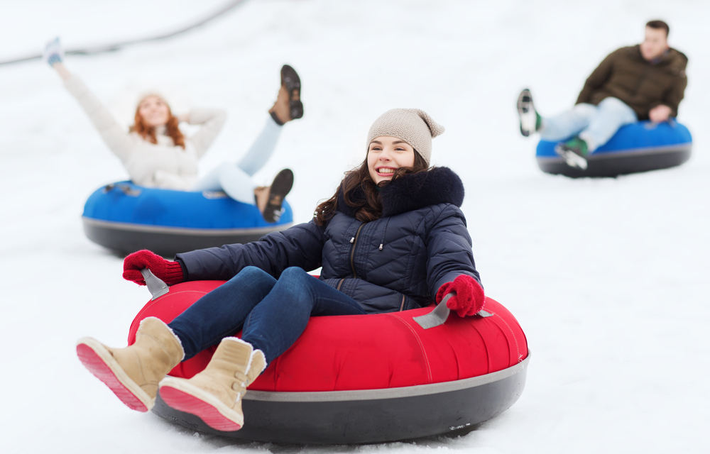 A photo of a girl smiling in a black coat and brown shoes that is snow tubing down a hill on a red snow tube with two friends behind her in blue snow tubes.