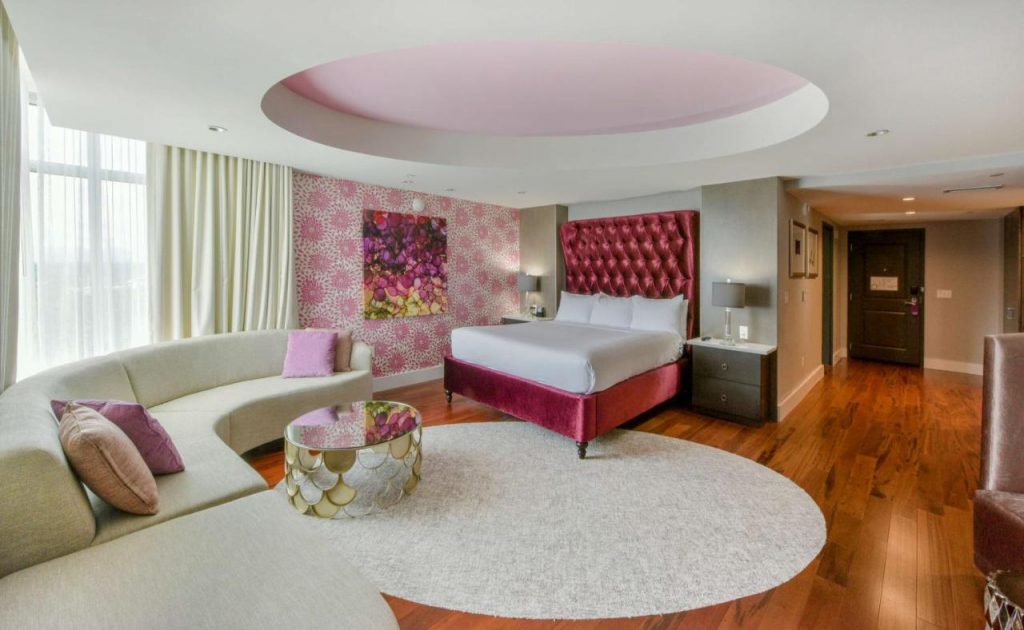 Photo of a suite inside the Hyatt Place featuring a pink velvet tufted bed frame, U shaped modern sofa, and colorful patterned wallpaper.