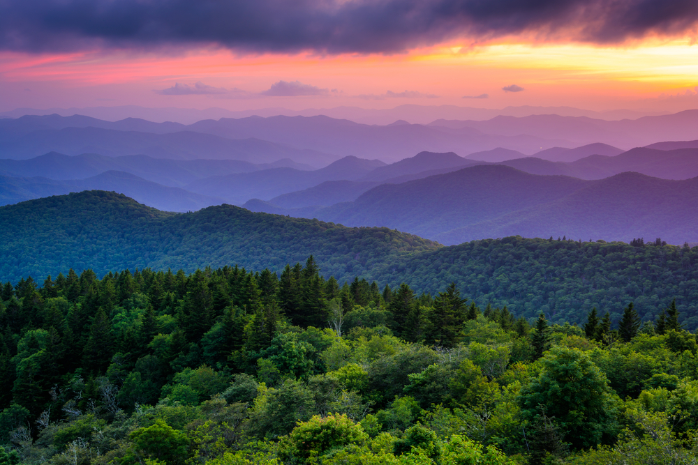 A view of the mountains and valleys in the Blue Ridge Parkway. The mountain tops have a blue tint to them in the distance and the sun is setting so the sky is purple, yellow, and orange. 