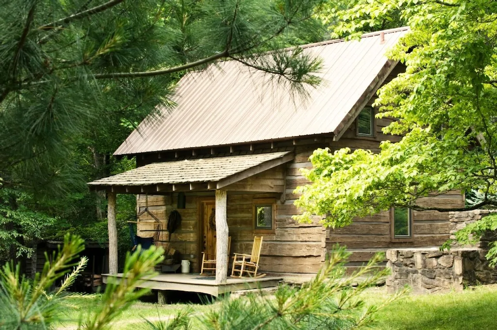 Photo of Lake Eden cabin, a small antique log cabin with rocking chairs on the front porch surrounded by trees. This cabin is where to stay in Asheville for nature!