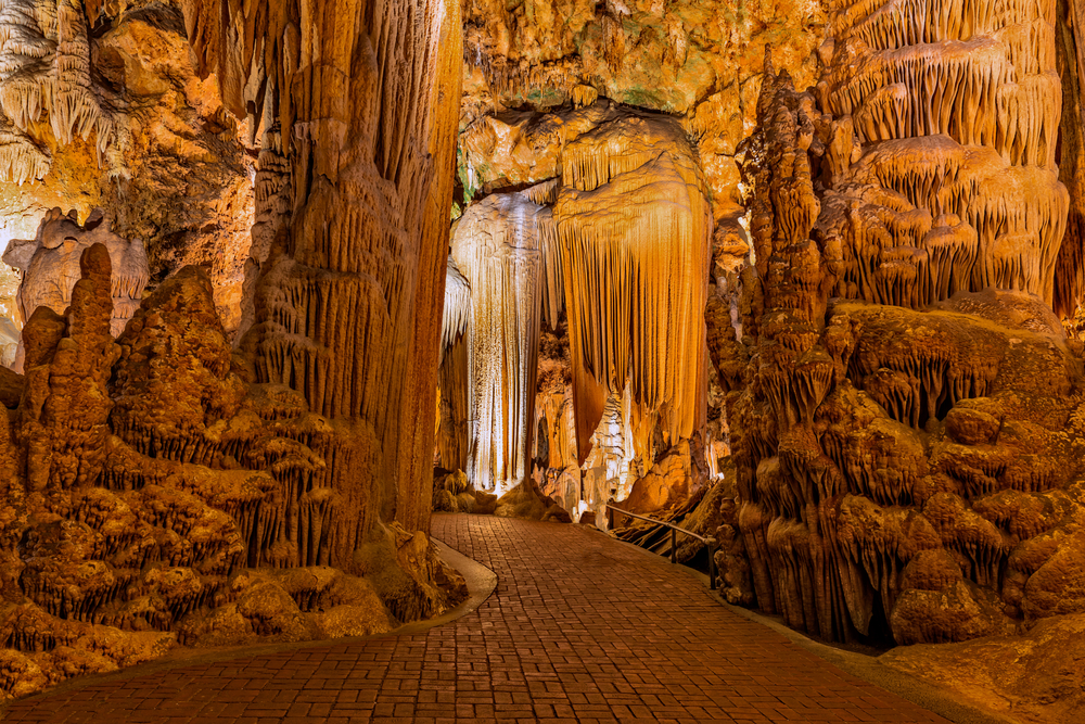 The inside of a massive cavern in Virginia. There are tall rock formations that make up the cavern in different shades of brown and yellow, and occasionally green. There is a brick walkway winding through the caverns.