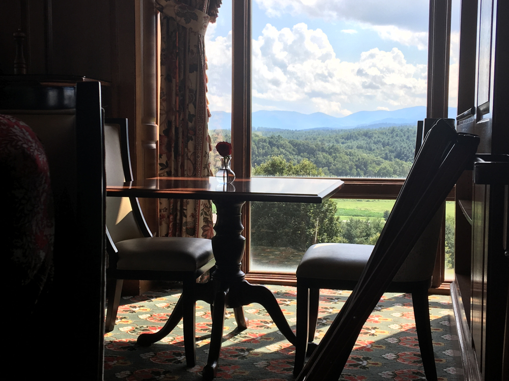 A small wooden table and chairs in an alcove that is next to a large floor to ceiling window. The window overlooks a view of the Blue Ridge Mountains and the Pisgah National Forest. It is one of the best views when visiting the Biltmore. 