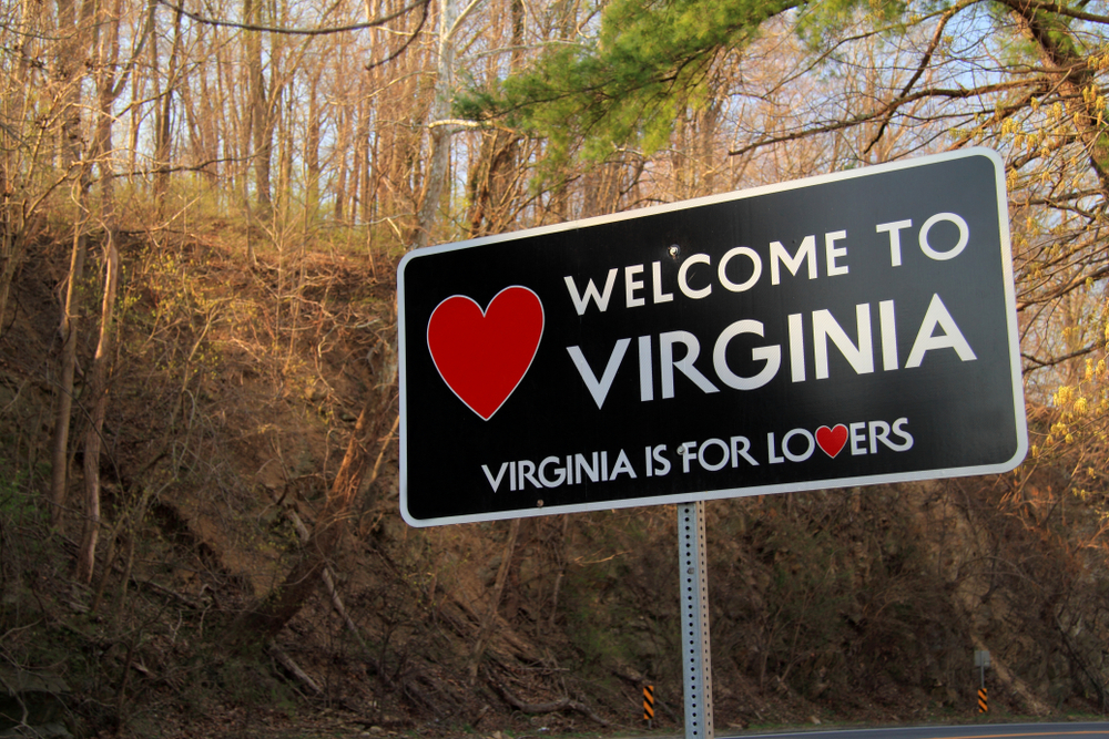 A black sign on the side of the road surrounded by trees. The sign has a red heart on it and says 'Welcome to Virginia. Virginia is for lovers'. The letters are white. 