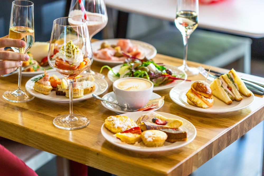 A fancy spread of brunch foods on a light wood table. You can see finger sandwiches, crepes, a salad, quiche, and a large wine glass with some sort of dessert with whipped cream in it. There are also two glasses with champagne. One of the delicious restaurants in Tupelo. 