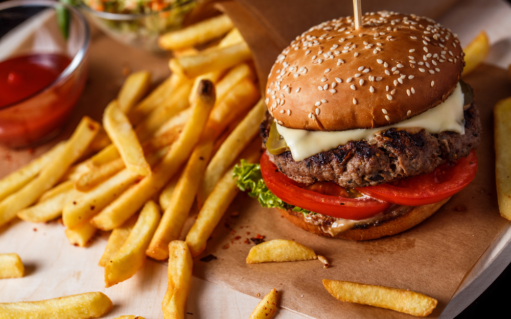 A gourmet burger on a piece of brown butcher paper on a table. The burger has cheese, tomato, lettuce, and pickles. All around the burger are fresh fries with some sort of spice on them and then a cup of ketchup. Best burger restaurants in Tupelo. 