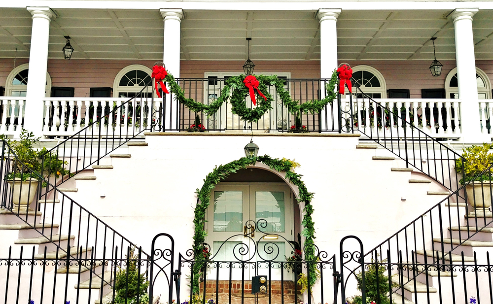 The front of a large white home with two large staircases on either side leading to a large front porch. There is greenery, a wreath, and red ribbons decorating the stair railing and door archways. 