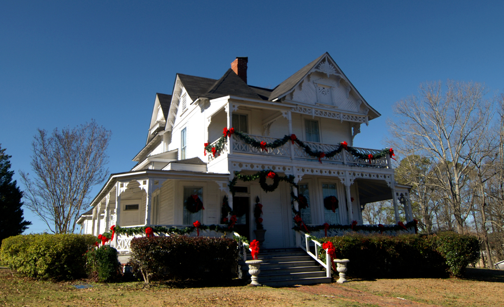 A large white historic Victorian farm house. It is decorated for Christmas with greenery on the railings of the porch, red ribbon, and wreathes. 