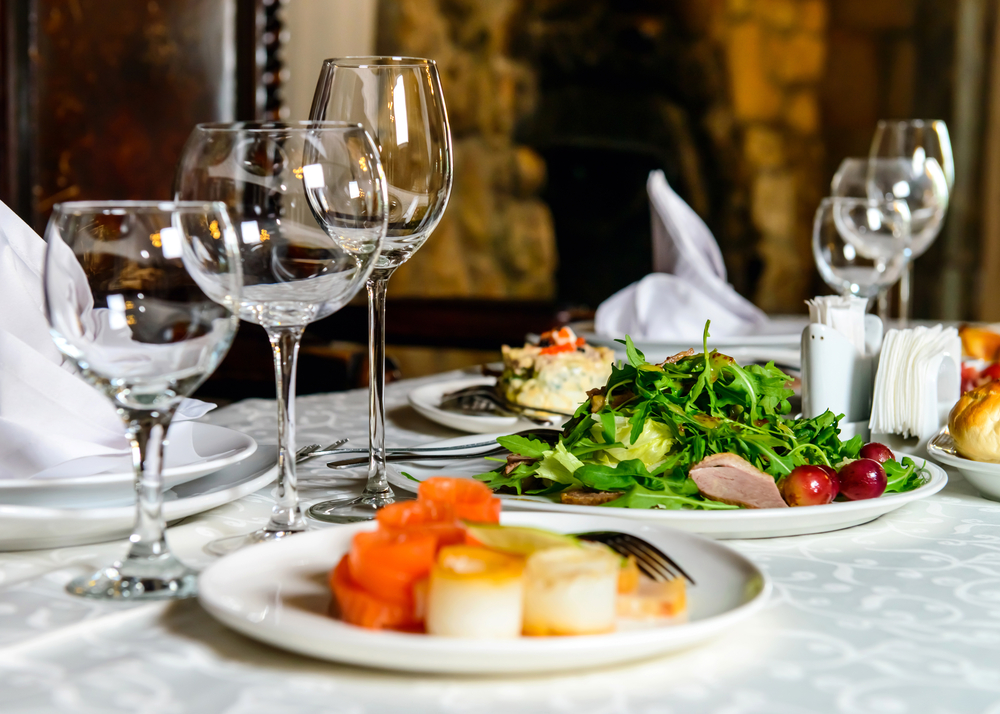 The table at one of the fine dining restaurants in Tupelo. There are empty wine glasses, napkins intricately folded, and a white tablecloth. There are plates with salad, rolls, quiche, and scallops, with other things. 