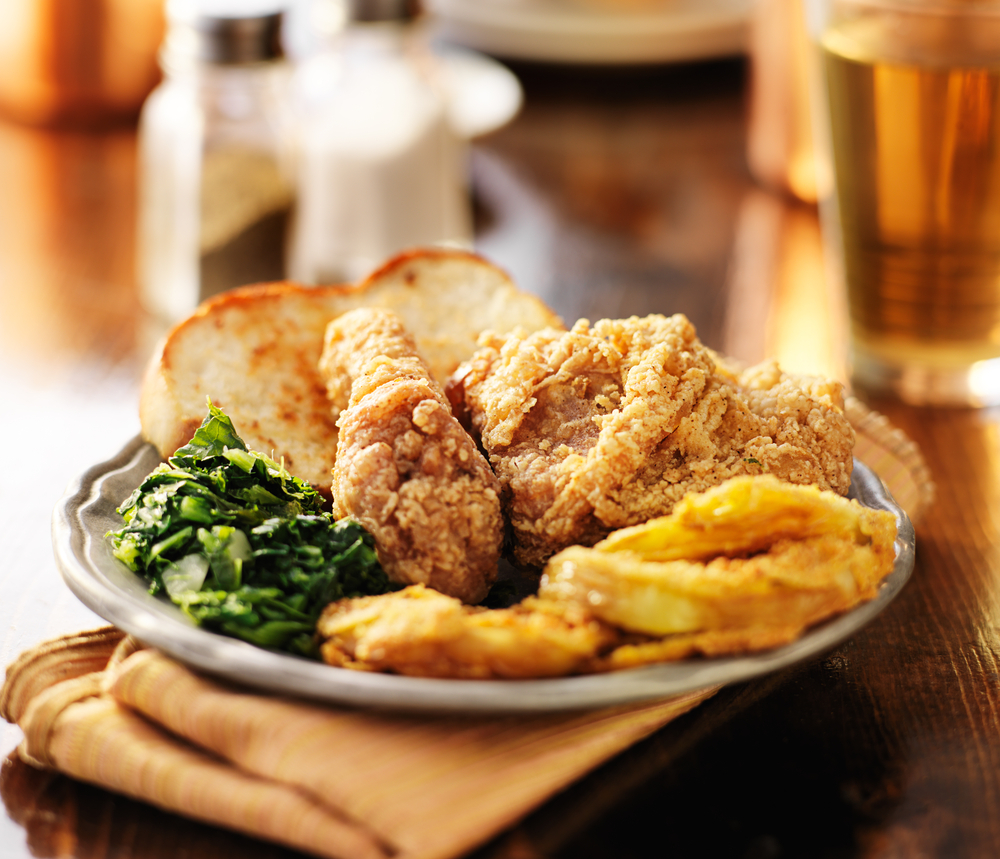 A plate of southern comfort food on a wooden table with a yellow napkin under it. The plate has a piece of toasted white bread, fried chicken, collards, and fried green tomatoes. 