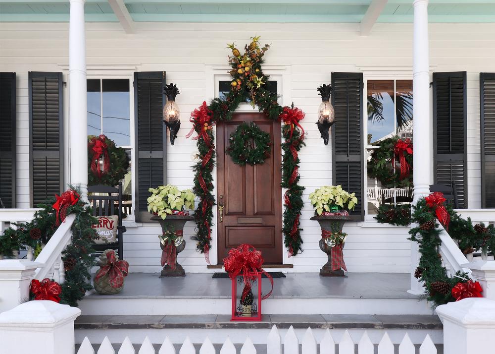 The front porch of a large white home that is elaborately decorated for Christmas with greenery, red ribbon, poinsettias, and other simple holiday decorations. 