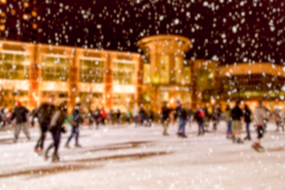 A bokeh image of people ice skating next to a large building. You can see snowflakes falling, the building appears to be orange in color, and there are lots of people on the ice. 