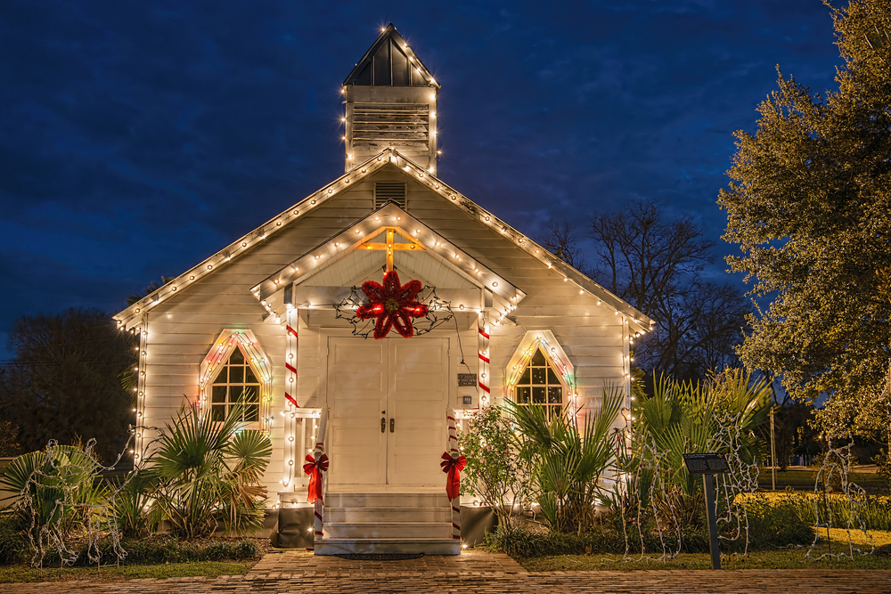 A simple old white church decorated with string lights, a red star, red ribbon, and a gold cross that lights up for celebrating Christmas in the south. In font of the church is palm trees. 