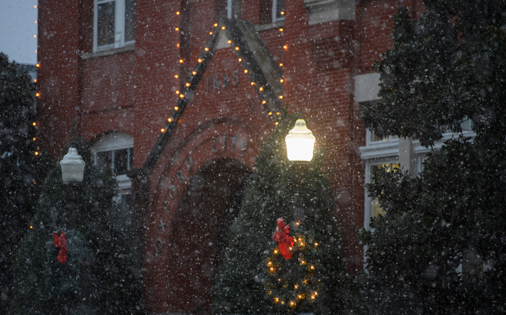 Snow falling in front of a large brick building in Oxford, Mississippi. The building is trimmed in large twinkle lights. There are also large evergreen trees and on the light poles there are wreaths lit up with red ribbon. 