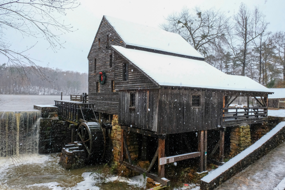 An old historic mill on the edge of a lake. It is snowing and there is snow on the ground. On the front of the old mill is a wreath with a red ribbon. 