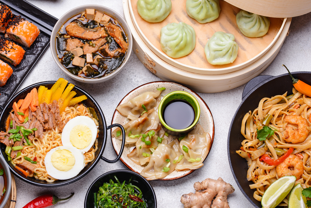 An assortment of Asian cuisine, featuring a steaming bowl of ramen with egg, vibrant sushi rolls, delicate green dumplings in a bamboo steamer, hearty noodle stir-fry with shrimp, dumplings with dipping sauce, and a refreshing seaweed salad, all arranged on a modern grey table setting.