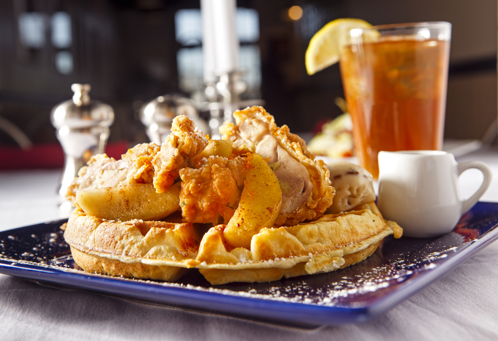 Crispy fried chicken on golden waffles, dusted with powdered sugar, accompanied by a side of syrup and a refreshing glass of iced tea with a lemon wedge, all served on a blue rimmed plate for a classic Southern comfort meal.





