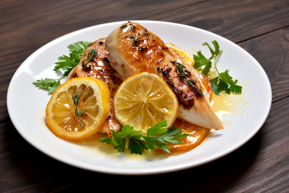 Grilled chicken breast garnished with fresh parsley and caramelized lemon slices, served on a white plate with a drizzle of light citrus sauce, presented on a dark wooden table.