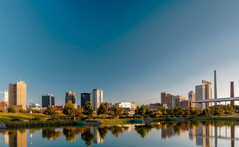 View of Birmingham city skyline reflected on a calm lake under a clear blue sky, showcasing a mix of modern and traditional architecture