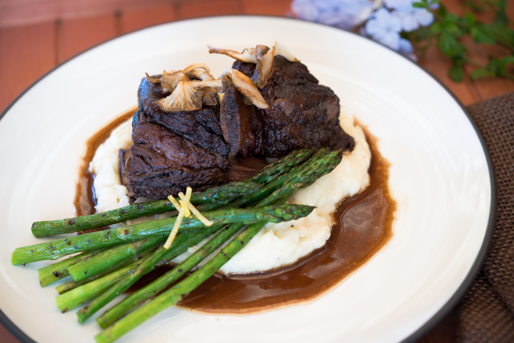 A succulent braised beef dish served over smooth mashed potatoes, garnished with golden-brown oyster mushrooms and a side of grilled asparagus, all drizzled with a rich brown sauce