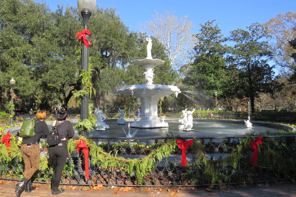 Two people standing against a railing looking at a large fountain in Savannah Georgia, one of the best places to spend Christmas in the south. The railing and light poles are decorated in fresh greenery and red ribbons. 