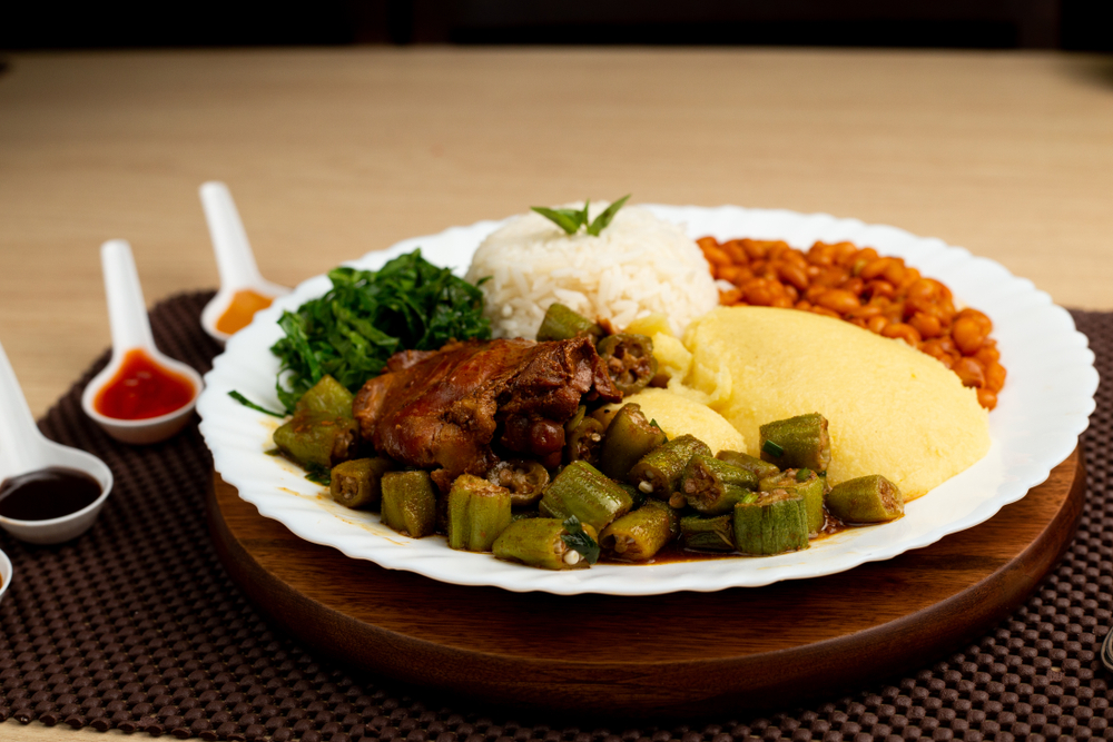 A white plate of soul food on a dark wood charger and brown woven placemat. On the plate there is okra, grits, rice, beans, collards, and some type of roasted meat. There are also spoons with sauce on them. One of the best restaurants in Tupelo. 