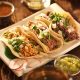 A plate with three different types of tacos on it. They are shredded chicken, shredded pork, and shredded beef. They have fresh cut cilantro and onion sprinkled on top. There is a lime wedge on the plate and dishes of different salsas on the wood table around the plate. A great meal to be found at restaurants in Austin.