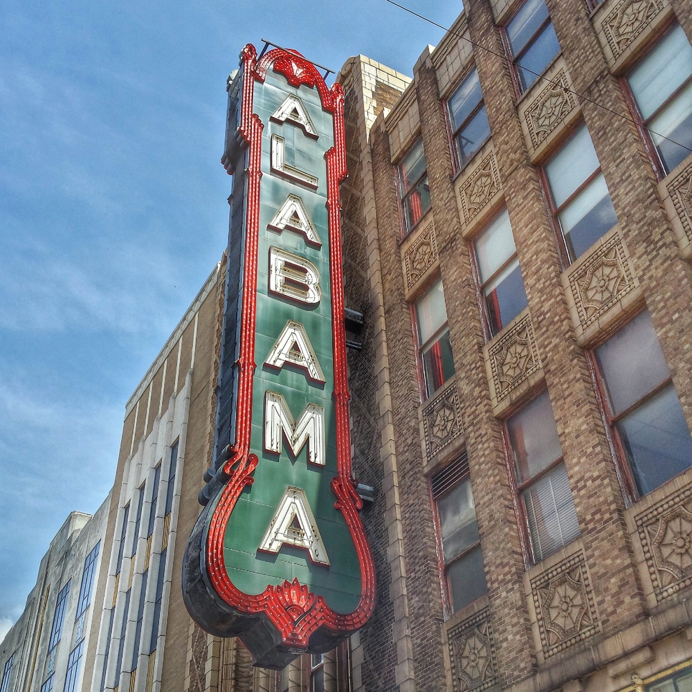 Close up picture of a sign reading "Alabama" for the Alabama Theatre, attatched to an old building with a blue sky in the background.