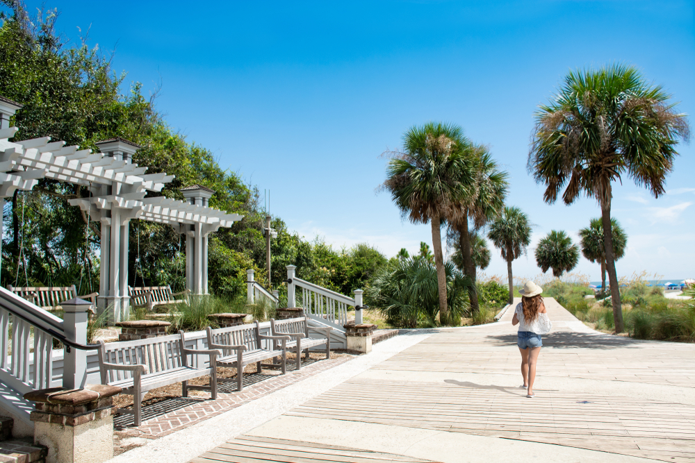 A woman with long hair, wearing a beach hat and shorts, walks past wooden benches and swings, away from the camera toward the beach, via a palm tree lined boardwalk at Coligny Beach Park.