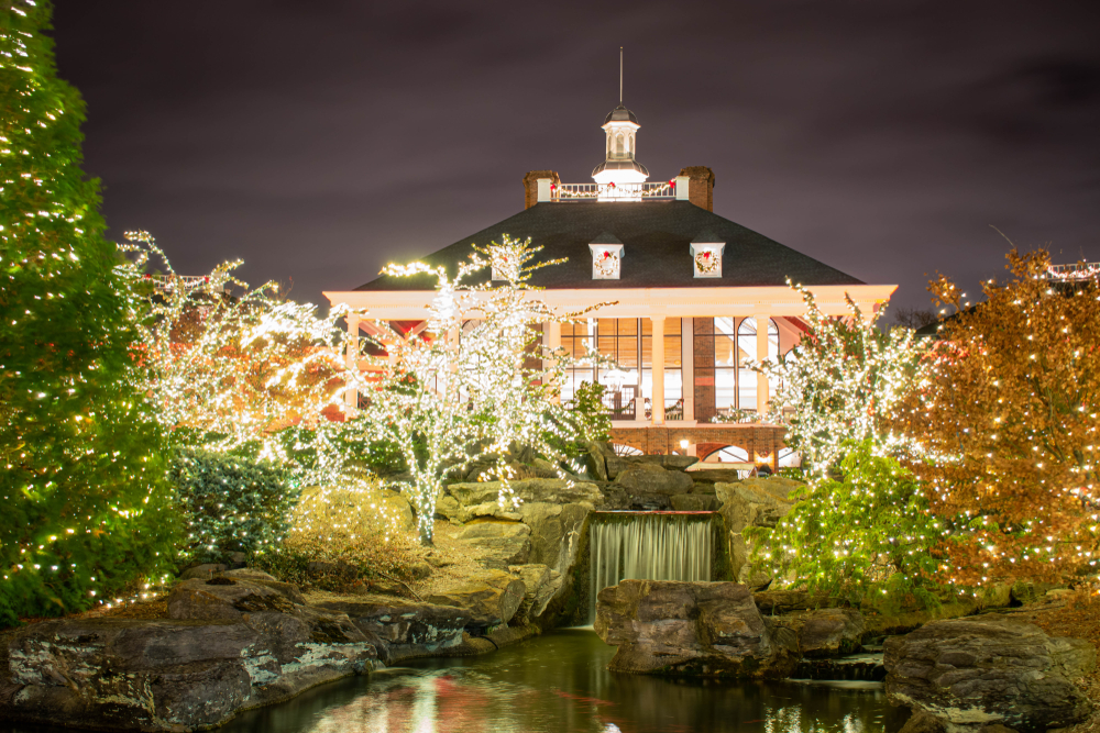 Opryland building that has a steeple in middle of roof. The building is surrounded by trees that are full of Christmas lights. Waterfall in front of the building.